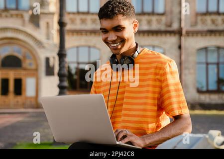 Junger schwarzer brünetter Mann mit Kopfhörern, der draußen am College ein Notebook benutzt Stockfoto