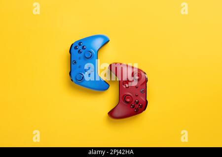 Zwei kabellose xbox Gamepads in Rot und Blau liegen hintereinander auf gelbem Hintergrund. Teamspiel, viel Platz Stockfoto