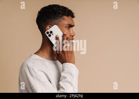 Profilporträt eines jungen, attraktiven, seriösen afrikaners, der telefoniert, während er über einem isolierten beigen Hintergrund steht Stockfoto