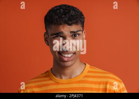 Porträt eines jungen, gut aussehenden, verwirrten afrikanischen Mannes mit Piercing, der in die Kamera schaut, während er über einem isolierten orangefarbenen Hintergrund steht Stockfoto