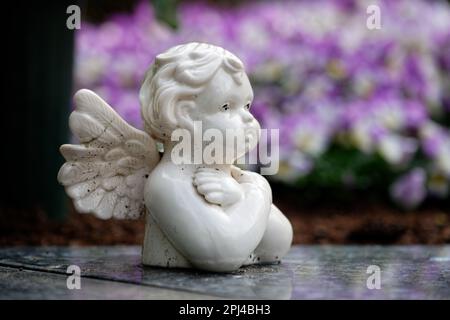 Ein kleiner, schmutzig weißer Putto auf einem Grabstein vor bunten Frühlingsblumen mit verschwommenem Hintergrund Stockfoto