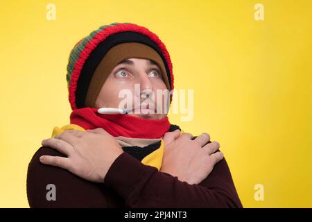 Ein junger Mann in warmer Kleidung auf gelbem Hintergrund hält ein elektronisches Thermometer in seinem Mund und ist von der hohen Temperatur schockiert Stockfoto