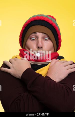 Ein junger Mann in warmer Kleidung auf gelbem Hintergrund hält ein elektronisches Thermometer in seinem Mund und ist von der hohen Temperatur schockiert Stockfoto