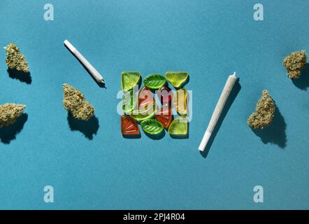Gummifrüchte liegen in einem Quadrat in der Mitte, trockene Knospen von medizinischem Marihuana und Gelenke liegen rechts und links. Cannabisbehandlung, auf einem blau-grünen B Stockfoto