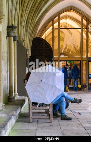 Salisbury, Wiltshire, Großbritannien, 31. März 2023: Menschen in den Kreuzgängen der Kathedrale von Salisbury mit Regenschirm bei nassem und windigem Wetter am letzten März. Stockfoto