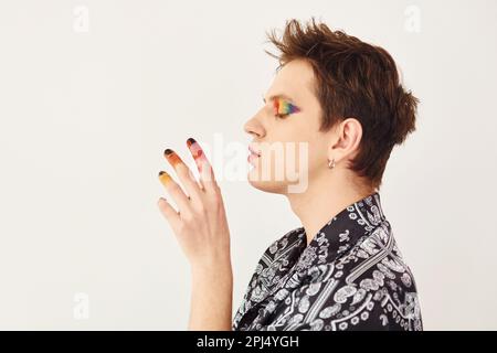 Ein junger schwuler Mann steht im Studio und posiert für eine Kamera. Mehrfarbiges Make-up auf Gesichtsfingern Stockfoto