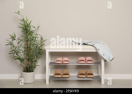 Elegante Aufbewahrungsbank mit verschiedenen Paar Gummipantoffeln und wunderschönen Zimmerpflanzen in der Nähe der weißen Wand im Flur Stockfoto
