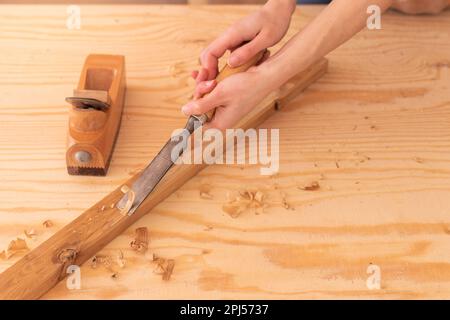 Draufsicht des Körperteils eines anonymen Zimmermanns, der Holz auf einer Planke mit Meißel auf einer Werkbank in der Nähe einer Hebebühne schneidet, während im Arbeitsraum mit Holz gearbeitet wird Stockfoto