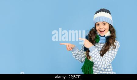 Stauned Kind mit lockigen Haaren in Hut und Schal. Teen Mädchen auf blauem Hintergrund. Porträt des Kindes trägt warme Kleidung. express positive Emotion. Winter fa Stockfoto