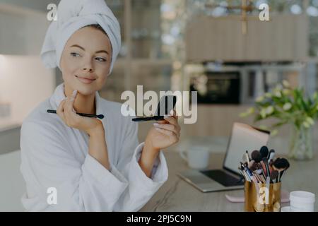 Zärtliche junge Frau hat morgendliche Routine auftragen tägliches Make-up hält Kosmetikbürste und Spiegel berührt sanft Gesicht trägt Bademantel und Handtuch auf Kopfhaltung Stockfoto