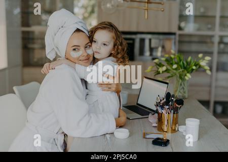 Eine attraktive Frau trägt Hydrogelpflaster unter den Augen, im Bademantel gekleidet, und ein eingewickeltes Handtuch umarmt ihre kleine Tochter mit Liebeshaltung zu Hause Stockfoto