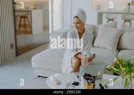 Entspannte, aufmerksame Frau in Bademantel und eingewickeltem Handtuch auf dem Kopf sitzt auf dem Sofa mit einer Tasse Getränk neben dem Tisch mit Kosmetikartikeln sieht pensi aus Stockfoto