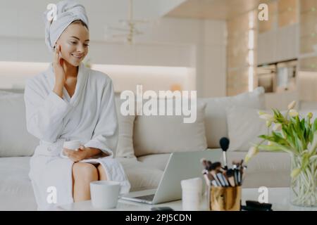 Foto einer lächelnden Frau trägt pflegende Creme auf das Gesicht auf Aussehen des Laptopbildschirms auf und gibt Tipps zur Hautpflege gekleidet in Bademantel und Handtuch sitzen bequem Stockfoto