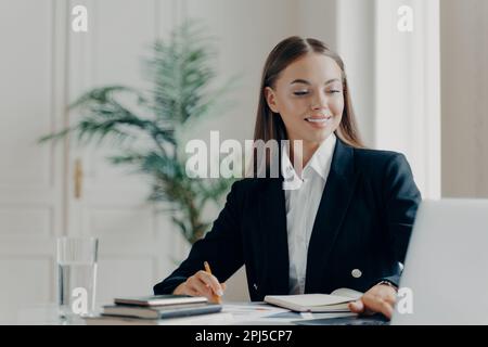 Portrait einer lächelnden jungen, weißen Geschäftsfrau im schwarzen Anzug, die am großen weißen Schreibtisch sitzt und mit dem Laptop arbeitet und mit Bl Sachen aufschreibt Stockfoto