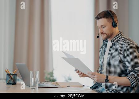 Seitenansicht eines lächelnden jungen Freiberuflers, der sich Dokumente ansieht und zufrieden ist, während er am Tisch sitzt und ein Notebook benutzt. Ein Typ mit Papieren und Stockfoto