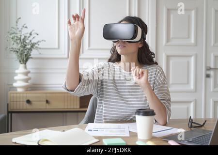 VR-App für die Arbeit. Junge fokussierte Frau mit Virtual-Reality-Headset, die 360-Grad-Inhalte ansieht und während des Sitzes in eine simulierte Umgebung eintaucht Stockfoto