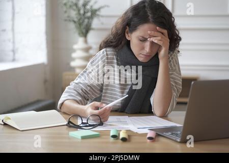 Junge lateinische Büroangestellte, die am Tisch sitzt und ein Digitalthermometer in der Hand hat, unter Kopfschmerzen und hohem Fieber bei der Arbeit leidet, weibliches Gefühl Stockfoto