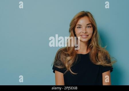 Foto einer reizenden, langhaarigen jungen Frau, die direkt in die Kamera schaut, mit einem zarten Gesichtsausdruck, gekleidet in einem legeren schwarzen T-Shirt, isoliert auf blauem Hintergrund Stockfoto