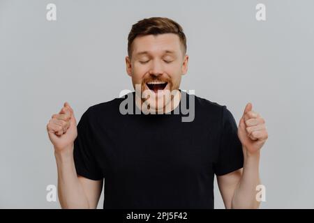 Wir feiern den Sieg. Junger, bärtiger Mann mit schwarzem T-Shirt, der fröhlichen, aufgeregten Ausdruck und positive Einstellung mit geballten Fäusten und offenem m zeigt Stockfoto