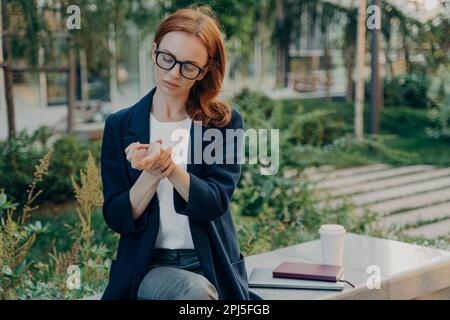 Unzufriedene Rothaarige leidet an Schmerzen im Handgelenk, nachdem sie im Stadtpark im Freien posiert. Sie ruht sich auf einer Bank aus. Sie trägt formelle Kleidung und berührt han Stockfoto