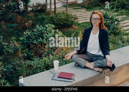 Friedliche, ruhige, rothaarige Geschäftsfrau, die Yoga mit geschlossenen Augen im Freien macht, sich in Lotus-Pose während abgelegener Arbeit im Park entspannt, Frauen, die Stress abbauen Stockfoto