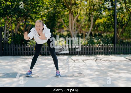 Outdoor-Aufnahme von zufriedenen gesunden sportlichen Frau neigt sich nach vorne hält die Handflächen angehoben macht Übungen in offenen Zügen im Park fühlt sich gut an trägt Sweatshirt Stockfoto