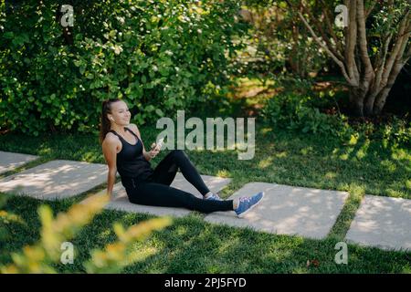 Seitenansicht einer erfrischten Frau macht nach dem aktiven Training Pause. Sie sitzt im Park und trägt Sportkleidung, und Turnschuhe hören Musik über Kabel Stockfoto