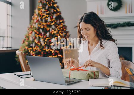 Wunderschöne italienische Frau, eine fröhliche Geschäftsfrau, die Weihnachtsgeschenke mit einem farbigen Band dekoriert, während sie am Schreibtisch im modernen Home Office gegen Backdro sitzt Stockfoto