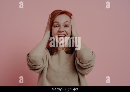 Ich kann meinen Augen nicht trauen! Überraschte junge, emotionale Frau mit rotem Haar, die den Kopf mit beiden Händen berührt und den Mund aufgeregt und angezogen hält Stockfoto