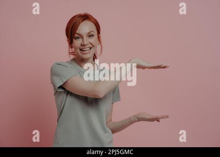 Junge, angenehme, rothaarige Frau im T-Shirt, die eine riesige Produktgröße oder etwas großes zeigt, mit Händen und lächelnd, während sie isoliert auf rosa Hintergrunnen steht Stockfoto