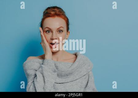 Porträt einer überraschten, rothaarigen Frau, die mit beeindruckendem Gesichtsausdruck in die Kamera schaut, sich glücklich fühlt, nachdem sie unerwartete Nachrichten gehört hat, isoliert Stockfoto