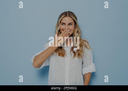 Eine glückliche blonde Frau, die mit der Hand den Mund schließt und die Überraschung von Freunden vorbereitet sieht, aufgeregte Frau, die in Erwartung auf etwas wunderbares lächelt, Stockfoto