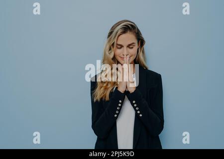 Hübsches, blondes Mädchen, das betet, und gefaltete Handflächen mit geschlossenen Augen hält, fleht und Wünsche wahr werden lässt, isoliert über blau Stockfoto