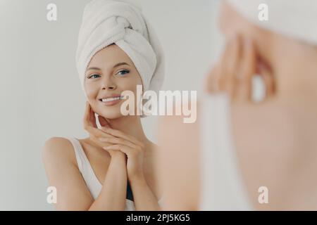 Wunderschöne junge Frau nach dem Duschen sieht in den Spiegel sanft berühren sanft weiche, gesunde, leuchtende Gesichtshaut, glückliche Millennial weiblich, die Schönheitsprodukte aufträgt Stockfoto
