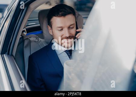 Junger positiver Geschäftsmann fährt im Auto auf dem Beifahrersitz in das Büro im Stadtzentrum, spricht mit fokussiertem Aussehen und lächelndem Smartphone. Seitenbild von Stockfoto