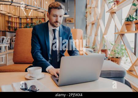 Junger selbstbewusster Geschäftsmann in formellen Anzügen arbeitet während der Kaffeepause online auf einem Laptop in der Lobby des Corporate Centers und bereitet die Präsentation für Geschäftsleute vor Stockfoto