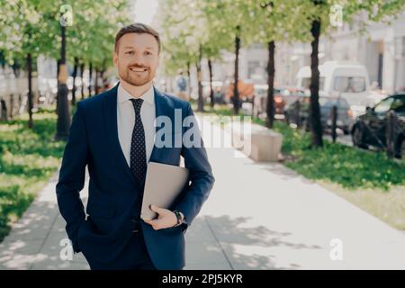 Entspannter, glücklicher, gutaussehender männlicher Büroangestellter in blauem Anzug hält Laptop mit wichtigen Daten und Präsentationen, macht einen Spaziergang im Park, während er eine Pause macht Stockfoto