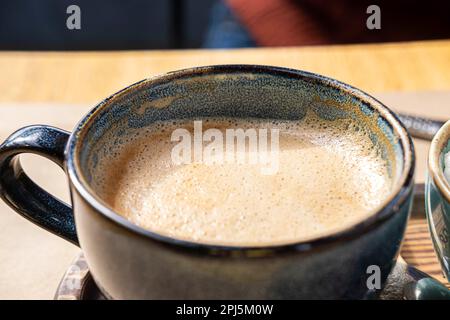 Blauer Keramikbecher mit heißem Cappuccino und Schaum auf einem hölzernen Café-Tisch. Wunderschöner Latte mit Schaumschaum. Trendiges, Authentisches, Modernes Porzellan-Set. Töpferei H Stockfoto