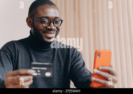 Lächelnder junger afroamerikanischer Geschäftsmann, der mit Kreditkarte und Smartphone am Arbeitsplatz in einem modernen Büro sitzt. Fröhlicher Mitarbeiter, der online arbeitet Stockfoto