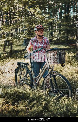 Aktive Frau, die freie Sommerferienzeit auf einer Fahrradtour in einem Wald verbringt. Frau trägt Fahrradhelm und Handschuhe halten Fahrrad mit Korb Stockfoto