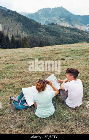 Familie, die eine Karte mit Bergversuchen durchsucht, auf dem Rasen sitzt und den Sommertag während des Urlaubs in den Bergen genießt. Menschen verbringen aktiv Zeit im Freien Stockfoto