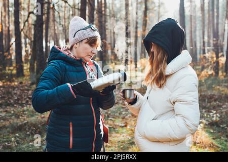 Mutter und Tochter mit Rucksack haben während der Herbstreise Pause und gossen am kalten Herbsttag ein heißes Getränk aus Thermoskanne. Aktive Frauen, die herkommen Stockfoto