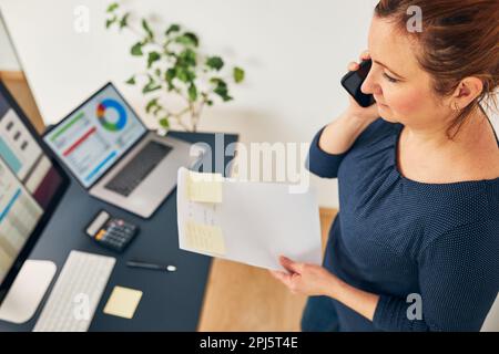 Unternehmerin, die ein Geschäftsgespräch mit dem Mobiltelefon führt. Geschäftsfrau, die im Büro arbeitet und Dokumente und ein Smartphone besitzt. Frau steht bereit Stockfoto