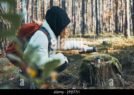 Frau, die während der Herbstreise eine Pause hat und am kalten Herbsttag ein heißes Getränk aus der Thermoskanne ausschenkt. Aktive Frauen, die in einem Wald wandern und aktiv Geld ausgeben Stockfoto