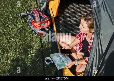 Frau, die die nächste Reise plant, während sie mit Karte im Zelt sitzt. Frau, die sich im Zelt während der Sommerferien beim Camping entspannt. Verbringen Sie aktiv Urlaub im Freien Stockfoto