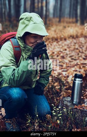 Frau mit Rucksack, die während der Herbstreise eine Pause hat und am kalten Herbsttag ein heißes Getränk aus einer Thermoskanne trinkt. Eine aktive Frau mittleren Alters, die um sich herumwandert Stockfoto