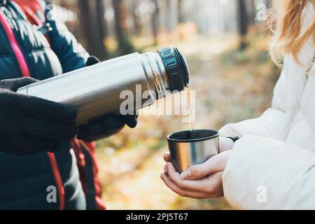 Frauen, die während der Herbstreise eine Pause haben und am kalten Herbsttag ein heißes Getränk aus der Thermoskanne ausschütten. Aktive Frauen, die in einem Wald wandern und aktiv Geld ausgeben Stockfoto
