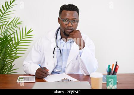 Nahaufnahme eines ernsthaften schwarzen Arztes, der an seinem Schreibtisch sitzt und ein Drehbuch schreibt Stockfoto