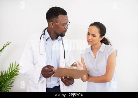 Schwarzer männlicher Arzt dokumentiert Symptome der jungen weiblichen Patientin beim GP-Besuch Stockfoto