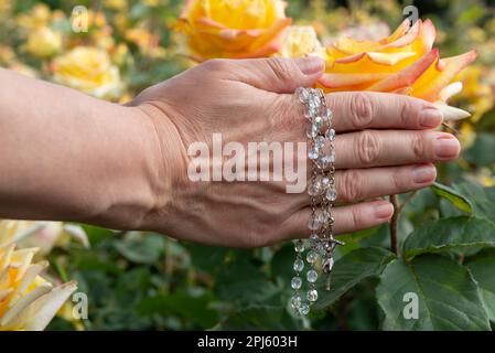 Nahaufnahme der Hände einer Frau mit einem Rosenkranz mit Glasperlen auf einem Hintergrund gelber Rosen, selektiver Fokus. Stockfoto